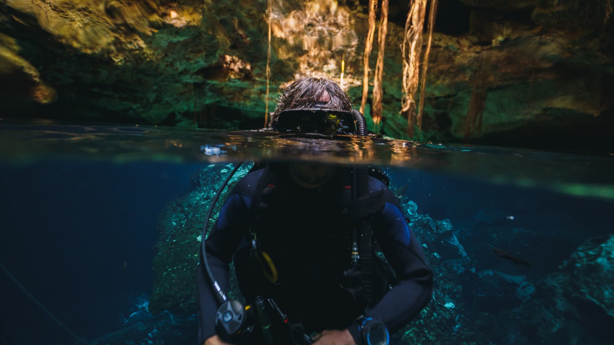 Split-level view of a diver at the surface of a cenote, jungle above and clear water below